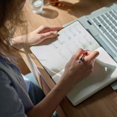 Woman at home journaling during a live online ancestral healing workshop, laptop open beside a family tree sketch